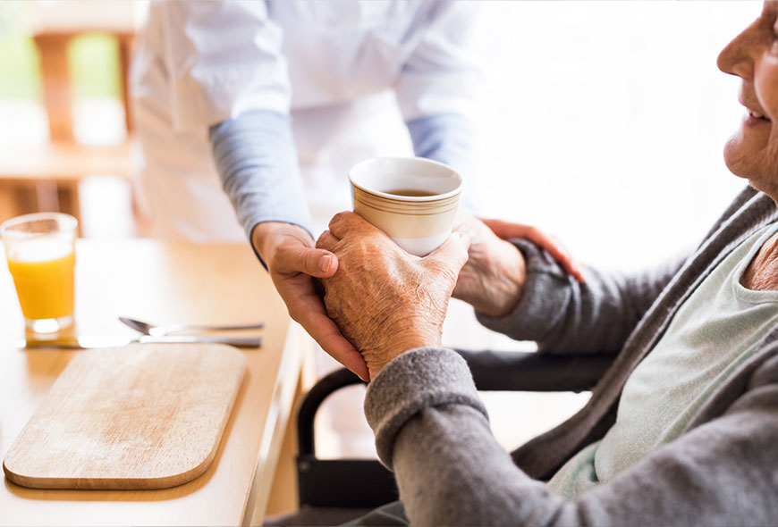 Eine Pflegekraft unterstützt mit seiner Hand die Hand einer älteren Dame, um ihr beim Kaffe trinken zu helfen.