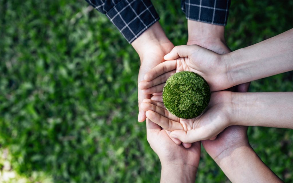 Hände mehrerer Personen tragen einen aus Gras angefertigten Globus, um den Planeten gemeinsam durch diverse nachhaltige, umweltfreundliche Maßnahmen zu schützen.